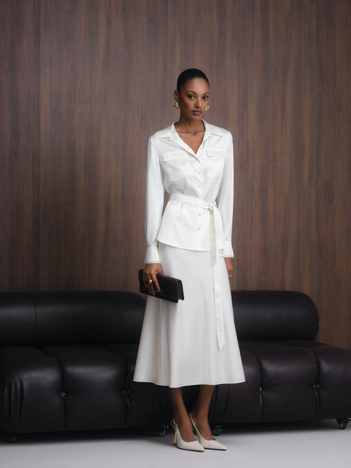 A woman models a white two-piece satin set consisting of a long-sleeve belted blouse and a matching midi skirt, standing in front of a dark wood-paneled wall and a black leather sofa.