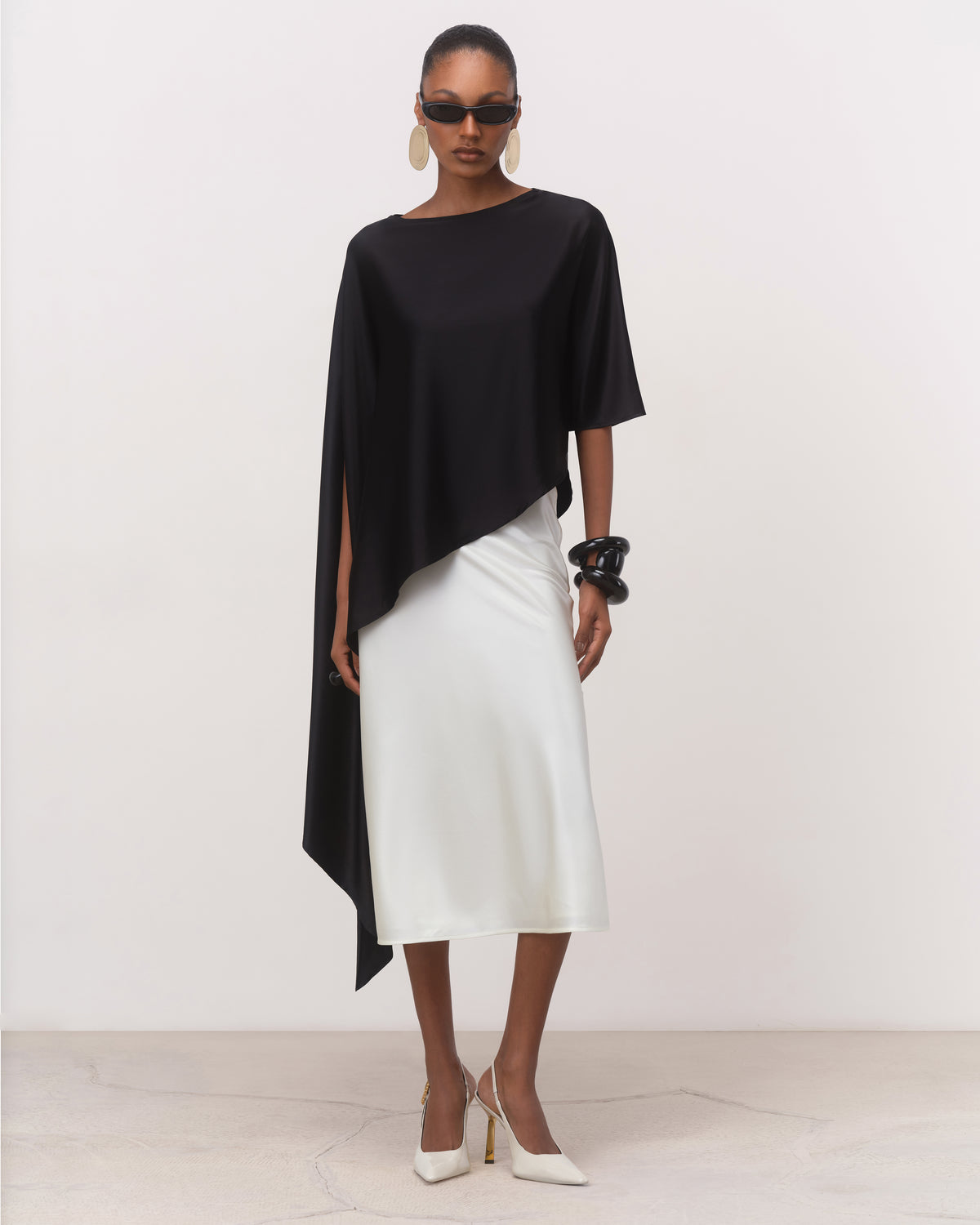 Anitas Muse black asymmetric silk cape top for women with boat neckline, worn with a white midi skirt, front view