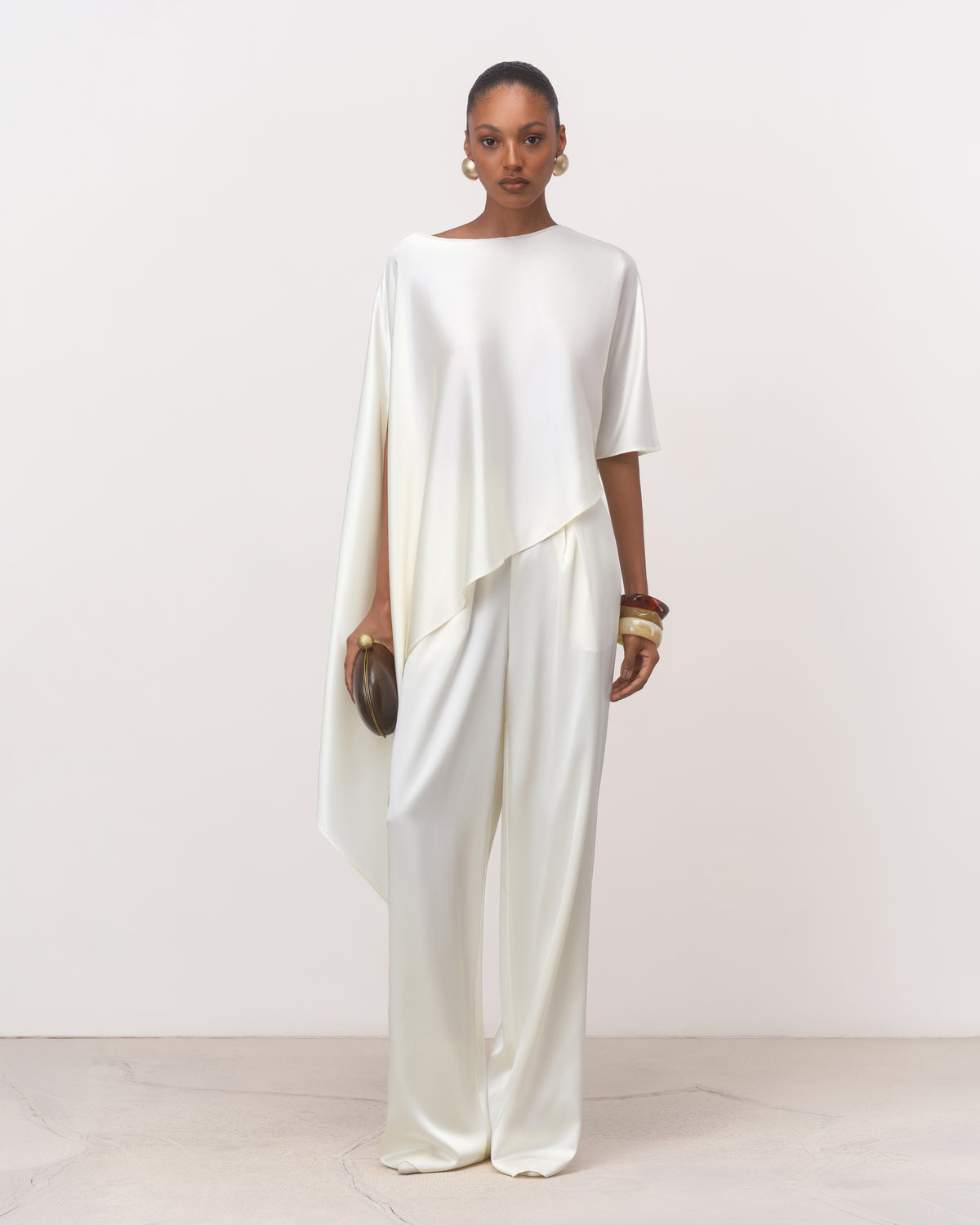 Anitas Muse pearl white asymmetric silk cape top for women with boat neckline, worn with white trousers, front view